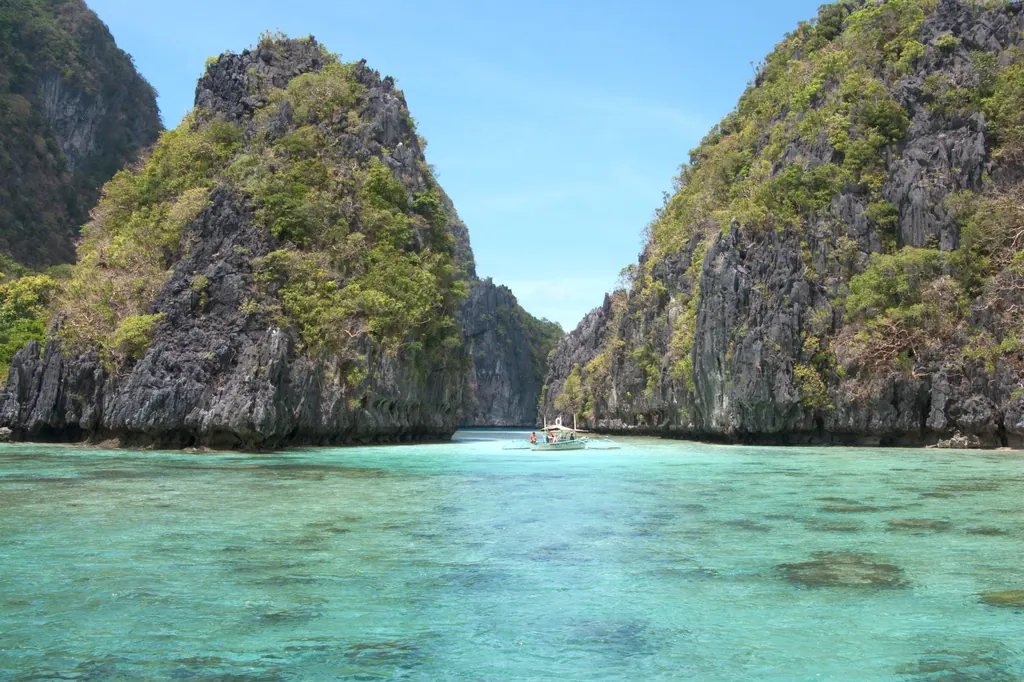 Turquoise lagoon between steep limestone cliffs in Bacuit Bay, El Nido, with an outrigger boat in the distance