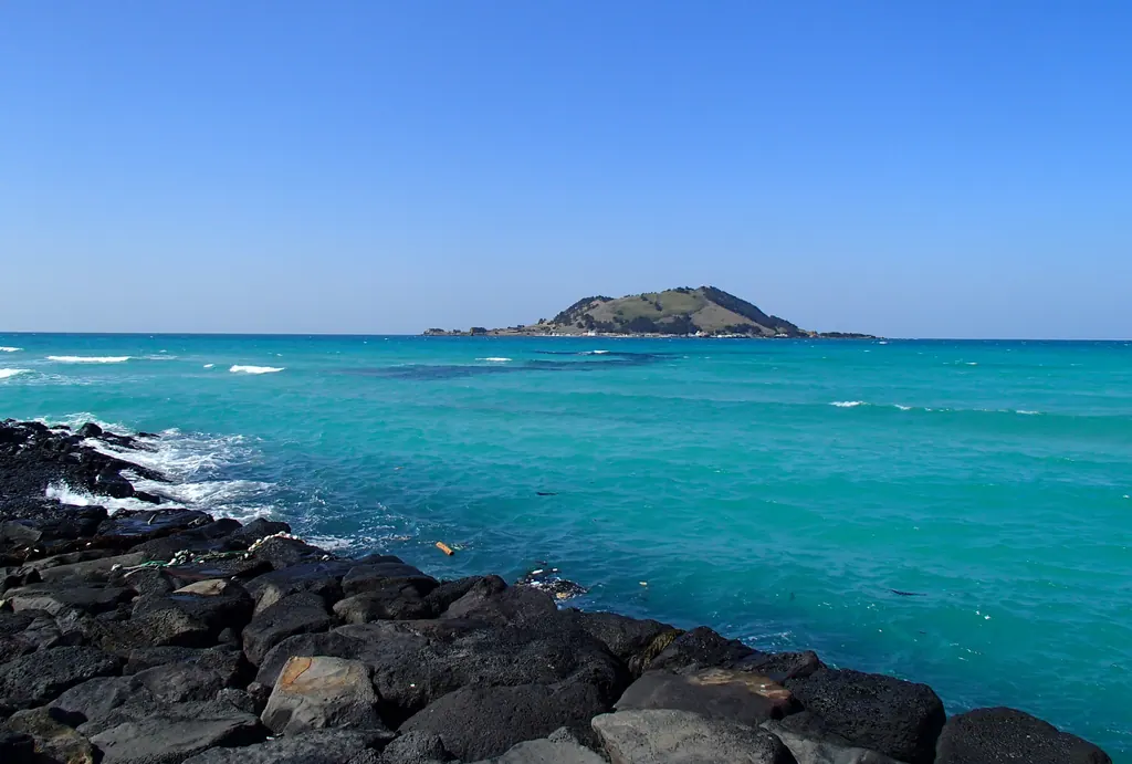 The turquoise water of Hyeopjae Beach on Jeju, with black lava rocks in the foreground and Biyangdo island offshore