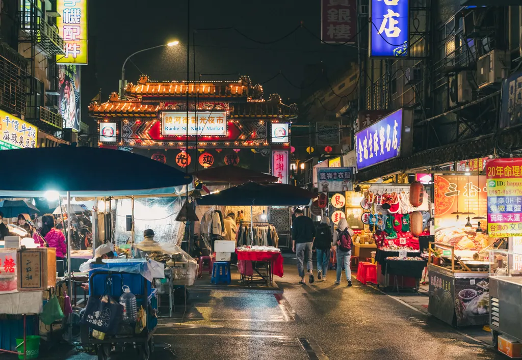Food stalls and neon signs on Raohe Street Night Market in Taipei, with the lit temple style entrance gate behind