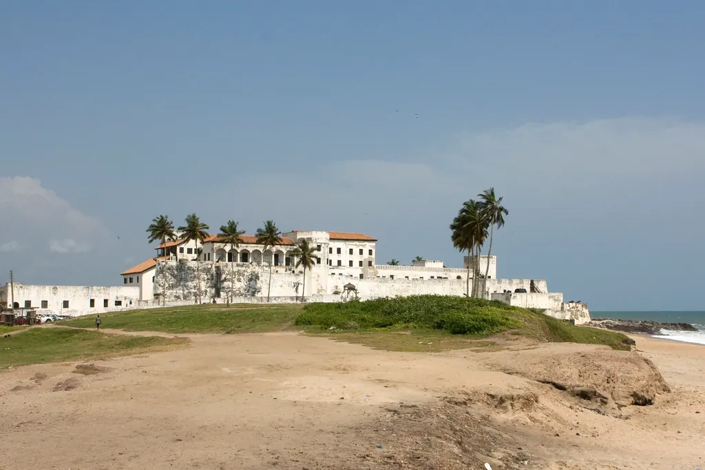 White walls of Elmina Castle on the coast of Ghana framed by palm trees under a blue sky