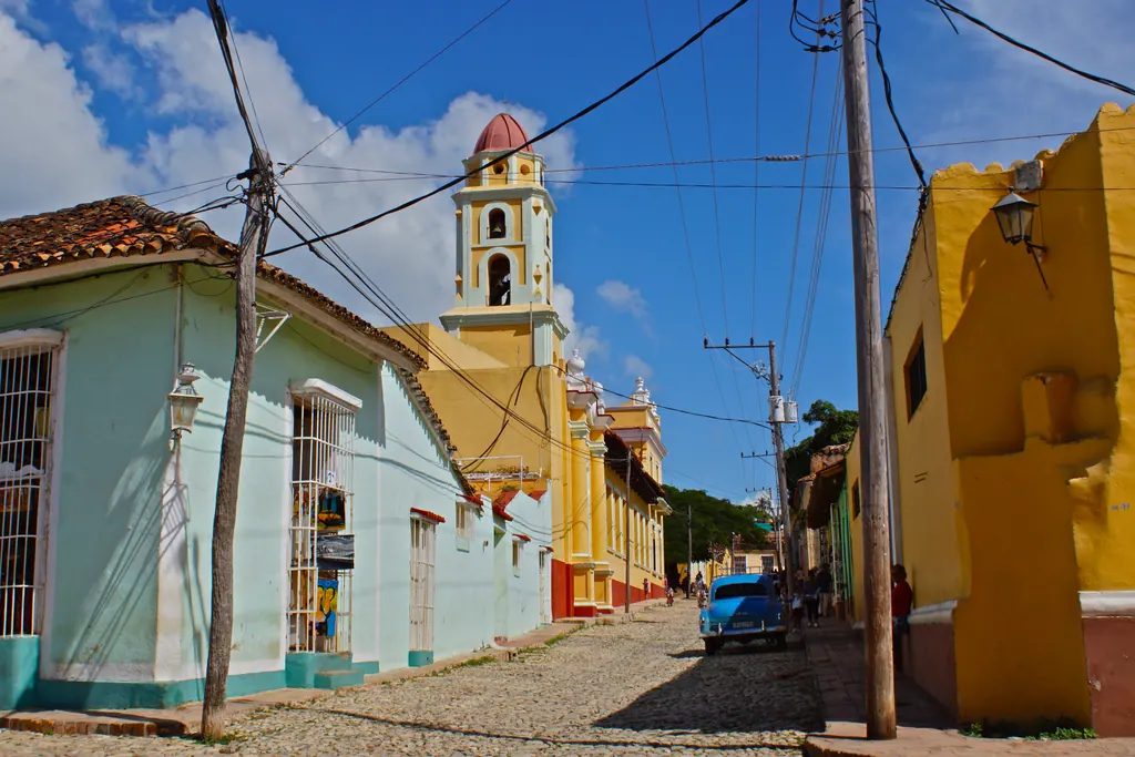 Cobblestone street in Trinidad, Cuba, lined with pastel colonial houses and a yellow church bell tower under a blue sky
