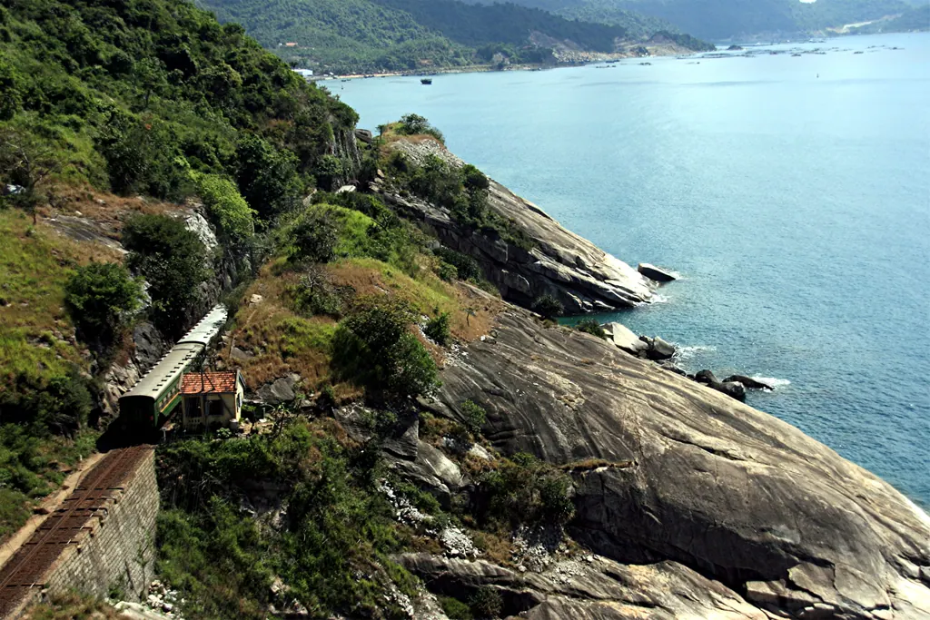 A green and cream passenger train runs along a rocky hillside above the turquoise sea on Vietnam's coastal railway