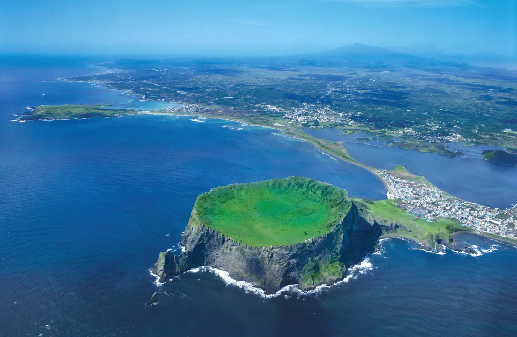 Aerial view of the green crater of Seongsan Ilchulbong rising from the blue sea on the east coast of Jeju Island
