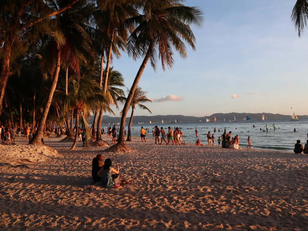 Palm trees and visitors on White Beach in Boracay at sunset, with sailboats on the water