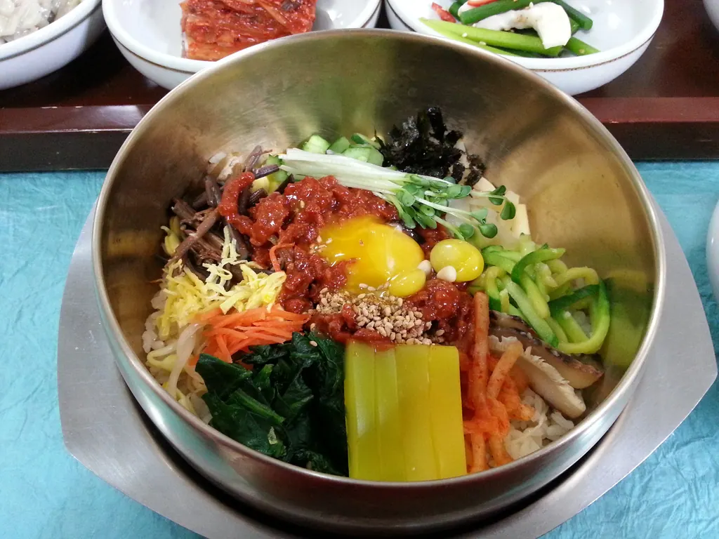 A brass bowl of bibimbap with vegetables, chili paste and a raw egg yolk, served with side dishes in Jeonju