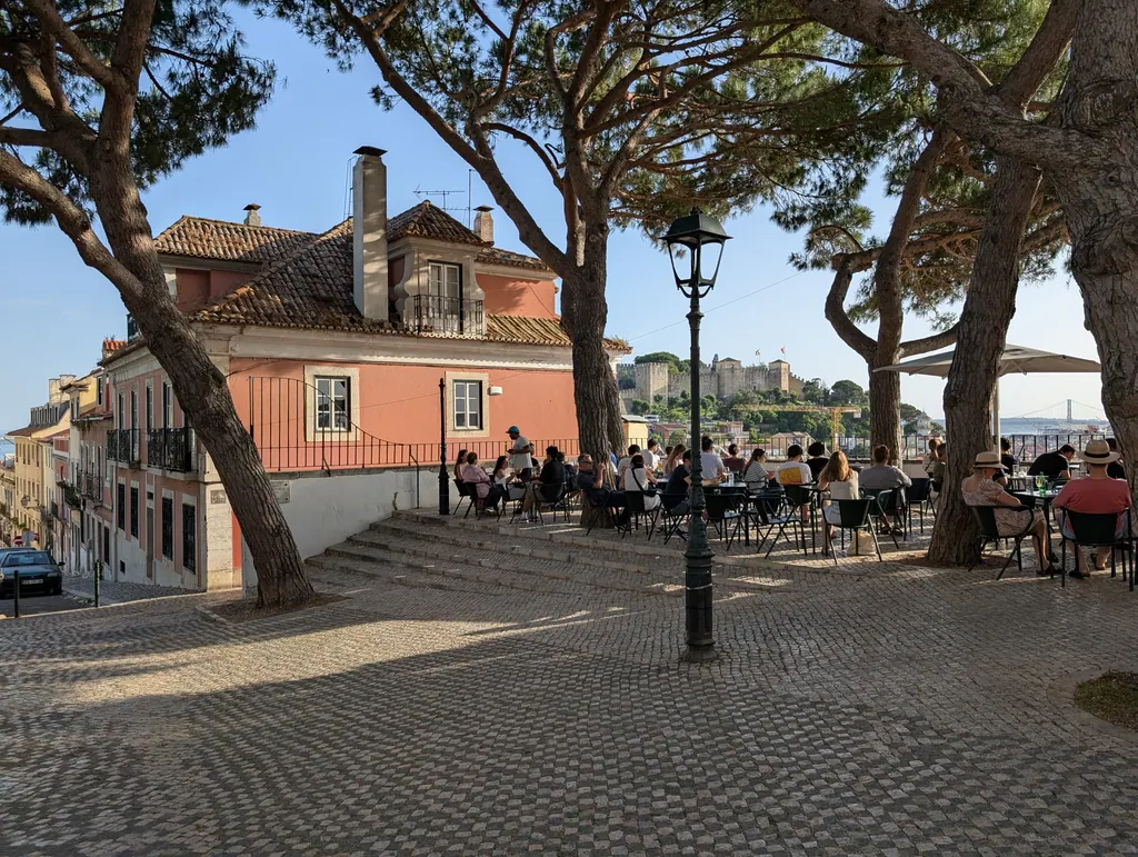 Outdoor restaurant terrace under pine trees at the Miradouro da Graça viewpoint in Lisbon on a sunny day